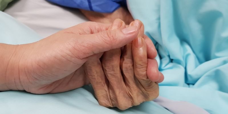 An image of a hand holding an elderly person's hand while they are in a hospital bed. Showing the need for palliative and end of life care.