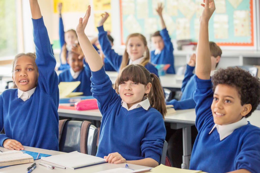 Primary school pupils with their hands raised in a classroom. The importance of epilepsy training in keeping pupils healthy and happy.